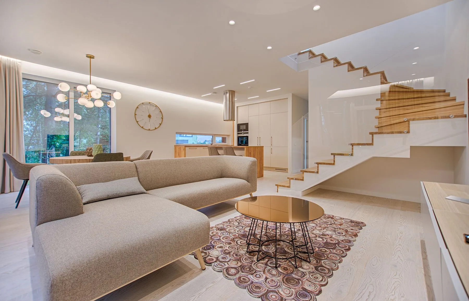 Salón abierto con sofá gris, escalera escultural de madera y cristal y cocina al fondo